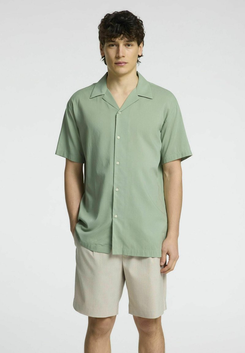 Camicia verde chiaro a maniche corte realizzata in tessuto morbido, con colletto e bottoni frontali, abbinata a pantaloni beige.