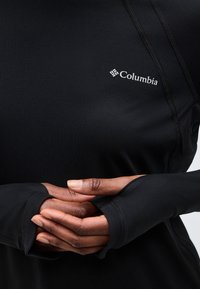 Χέρια διπλωμένα πάνω σε μαύρο αθλητικό μπλουζάκι μακρυμάνικο με τρύπες για τους αντίχειρες και λευκό λογότυπο Columbia στο στήθος.