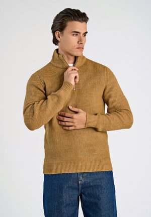 Maglione lavorato a maglia di colore giallo senape con colletto alto e chiusura con zip. Presenta una texture a coste e maniche lunghe, abbinato a jeans di denim.
