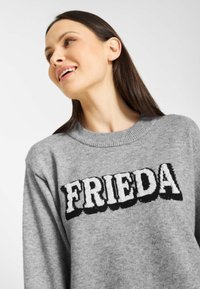 Szary sweter z dzianiny z okrągłym dekoltem i wyraźnie wyeksponowanym napisem "FRIEDA" w teksturowanej czarnej czcionce.