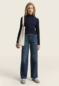 Maglia a maniche lunghe blu navy abbinata a jeans blu a gamba larga. La modella tiene una borsa tote color crema. Calzature: sneaker azzurre con suola marrone.