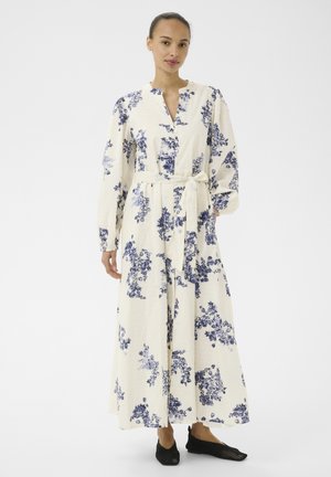 Ženska, oblečena v dolgo belo obleko z modrim cvetličnim vzorcem, z zavezanim pasom, dolgimi rokavi, obuta v črni čevlji brez vezalk, stoji pred belim ozadjem.