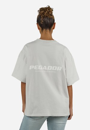 Kvinde med krøllet hår sat op i knold iført en lys grå oversized t-shirt med teksten "PEGADOR" på ryggen og lyseblå jeans, set bagfra.