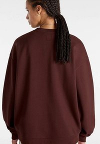 Vinröd oversized sweatshirt med rund halsringning, sänkta axlar och ribbade ärmslut, med en kvadratisk etikett på baksidan. Slät textur.
