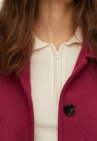 Chaqueta texturizada burdeos con cuello en muesca, con cierre de botón negro. Debajo, un top crema acanalado con cuello con cremallera.