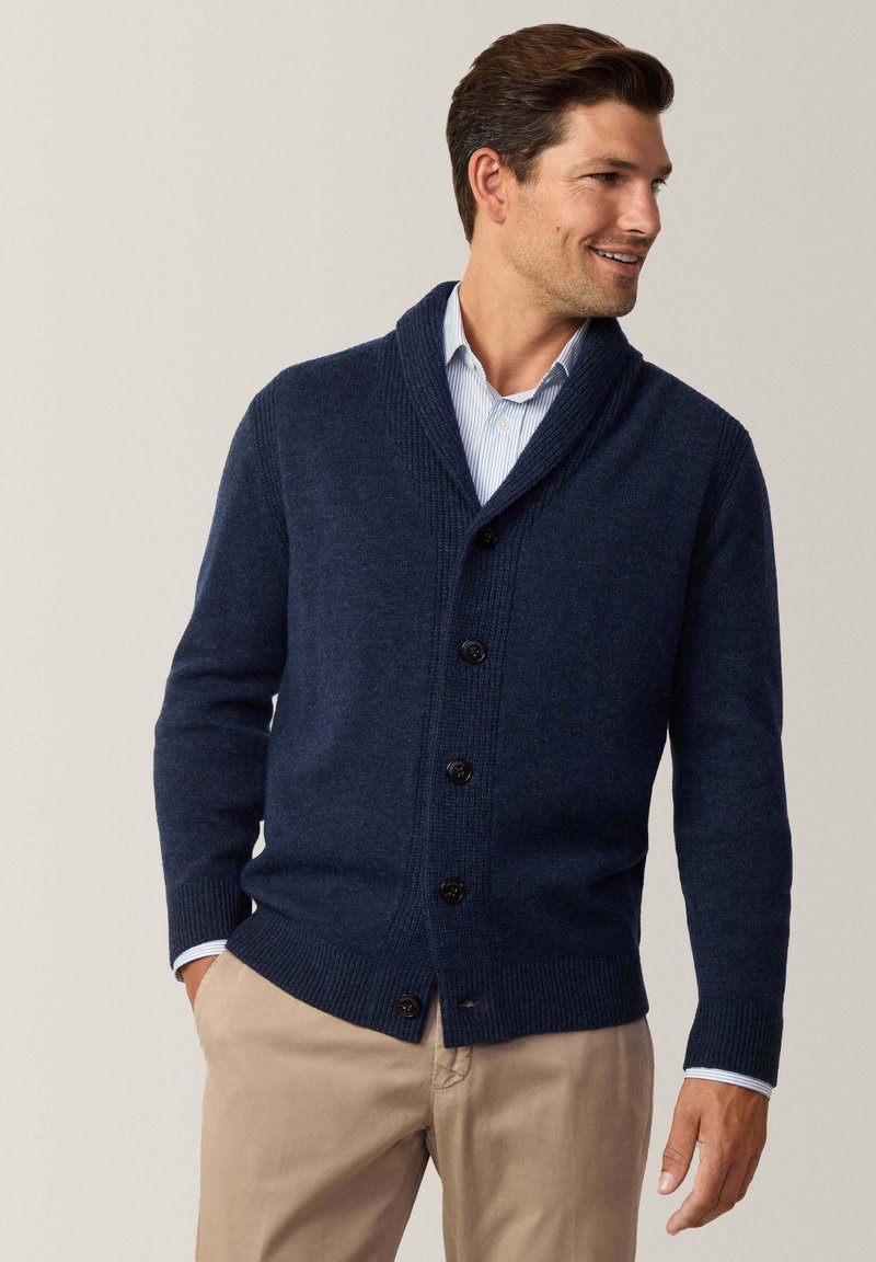 Cardigan lavorato a maglia color navy con collo a scialle, polsini e orlo a coste, sei bottoni neri, indossato sopra una camicia azzurra con colletto.