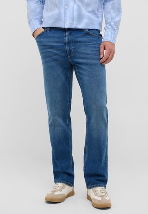 Mand iført blå jeans, lyseblå ternet skjorte, hvide og beige sneakers, stående med hænder delvist i lommerne på ensfarvet baggrund.