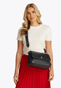 Žena s dlouhými blond vlasy, která má na sobě bílý krátký rukáv top, červenou plisovanou sukni a černou kabelku přes rameno se širokým popruhem.