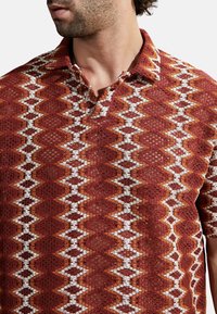 Polo de manga corta en óxido, crema y naranja con un patrón geométrico. La tela presenta un diseño texturizado y de tejido abierto, así como un cuello.