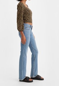 Top corto floral con mangas largas abullonadas, base negra, flores amarillas; vaqueros de tiro alto en azul claro; sandalias negras de esc slide con tiras gruesas.