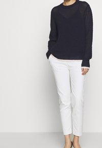 Person in einem strukturierten, navyblauen Langarmpullover über einem hellrosa Top, weißen Slim-Fit-Hosen und schwarzen Schuhen, steht mit den Händen in den Taschen.
