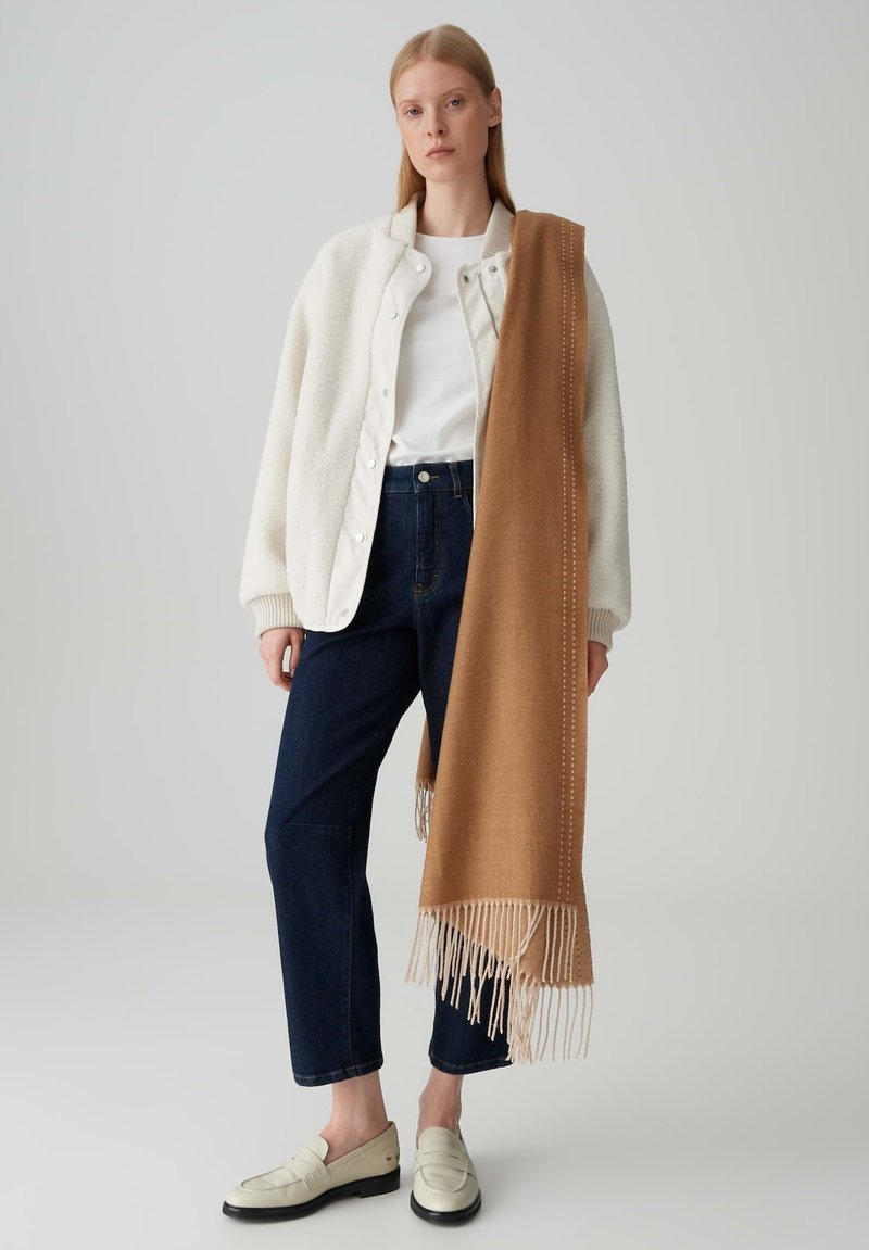 Giacca in pile bianca con bottoni, camicia bianca semplice, jeans dritti blu scuro, sciarpa beige con frange e mocassini crema.