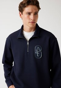 Marine blauwe half-zip pullover van textuurstof, met een geborduurd 'G' logo in lichtblauw en wit, voorzien van een ritskraag.