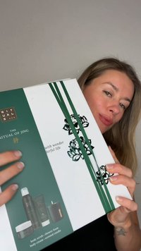 Caja blanca con detalles verdes que presenta un diseño floral, una cinta y productos: exfoliante corporal, espuma de ducha, crema corporal y vela aromática.