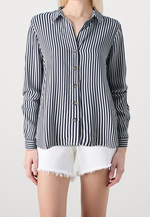 Woman wearing a long-sleeve vertical black and white striped button-up shirt with white frayed denim shorts.