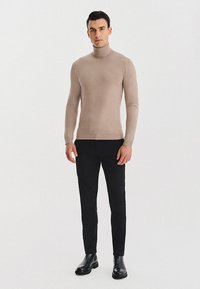 Beżowy sweter z golfem o dopasowanym kroju, wykonany z gładkiego dzianinowego materiału, zestawiony z czarnymi wąskimi spodniami i czarnymi skórzanymi butami.