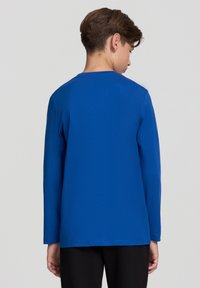 Koszula z długim rękawem w jasnoniebieskim materiale, z okrągłym dekoltem i gładką teksturą. Widok z tyłu pokazuje prosty, nieozdobiony styl.