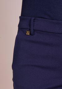 Pantalons bleu marine avec une texture lisse, ornés d'un accent logo doré sur la taille et d'un design à plat sur le devant.