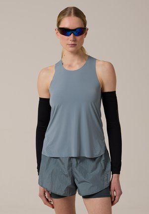 Mujer con camiseta deportiva gris sin mangas, pantalones cortos a juego, manguitos negros y gafas de sol deportivas reflectantes, posando frente a un fondo neutro.