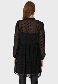 Schwarz gepunktetes Kleid mit langen, durchsichtigen Ärmeln, einem taillierten Oberteil und einem ausgestellten Rock. Die Rückansicht zeigt einen strukturierten, leichten Stoff.