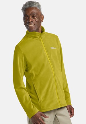 Homme âgé souriant aux cheveux gris portant une veste polaire zippée vert citron et un pantalon beige, debout devant un fond uni.