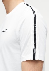 Λευκό βαμβακερό T-shirt με στρογγυλή λαιμόκοψη, κοντά μανίκια και μαύρες πλευρικές λεπτομέρειες από δίχτυ, με ταινία με λογότυπο στους ώμους.