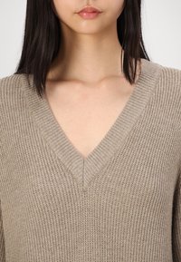 Beige stickad tröja med V-halsdesign och ribbad textur. Mjuk material med en avslappnad passform. Inga synliga mönster eller detaljer av hårdvara.