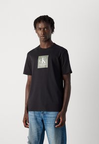 Joven con trenzas cortas que lleva una camiseta negra de Calvin Klein y jeans de color azul claro, de pie frente a un fondo blanco liso.
