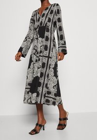 Robe maxi noire avec imprimé paisley blanc, décolleté en V, manches longues et silhouette décontractée. Associée à des talons noirs à lanières.