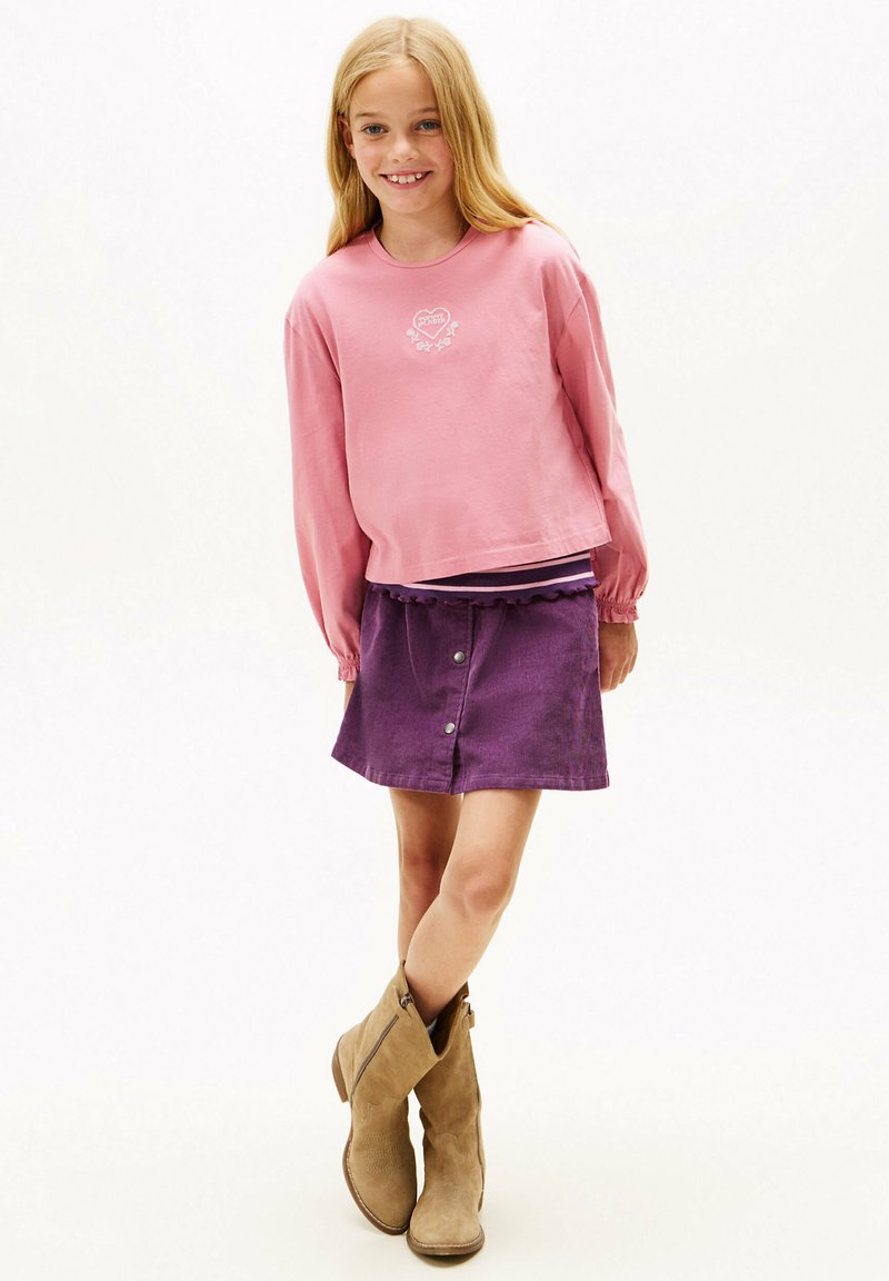 Różowa koszula z długim rękawem z graficznym sercem, nałożona na fioletową spódnicę z guzikami, zestawiona z beżowymi botkami za kostkę. Gładkie faktury tkaniny.