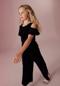 Zwart fluwelen off-shoulder jumpsuit met gerimpelde halslijn, met een aansluitend lijfje en wijde pijpen. Gadrd met zilveren glittersandalen.