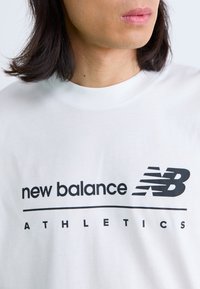 T-shirt en coton blanc présentant "new balance" et "ATHLETICS" imprimés en noir. Le logo comprend un 'NB' stylisé avec des lignes.