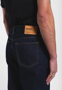 Tummansiniset denimfarkut, joissa näkyvät keltaiset saumat. Takana suorakaiteen muotoinen nahkapalainen, jossa brändäys ja hoito-ohjeet.