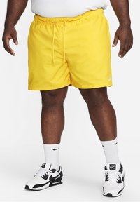 Shorts de bain jaunes avec une taille élastique et un cordon de serrage, présentant un petit logo Nike blanc. Portés avec des baskets Nike noir et blanc.