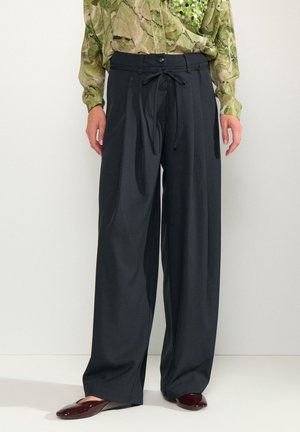 Personne portant un pantalon large noir taille haute avec un lien et un bouton à l'avant, associé à un haut à manches longues vert à motifs et des chaussures plates foncées.