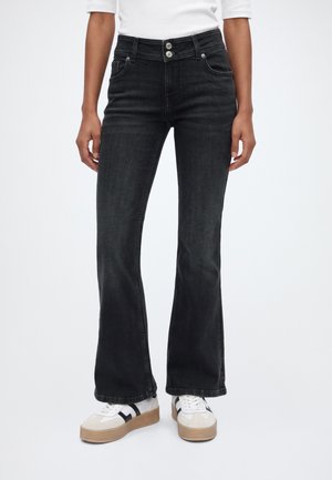 Person iført sorte højtaljede vide jeans med dobbelt knaplukning og hvide sneakers med sorte striber på en ens hvid baggrund.