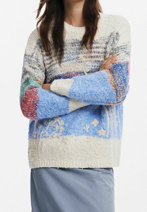 Kvinde med langt brunt hår iført en flerfarvet, tekstureret strikket sweater med blomsterprint og en lyseblå nederdel, armene krydset.