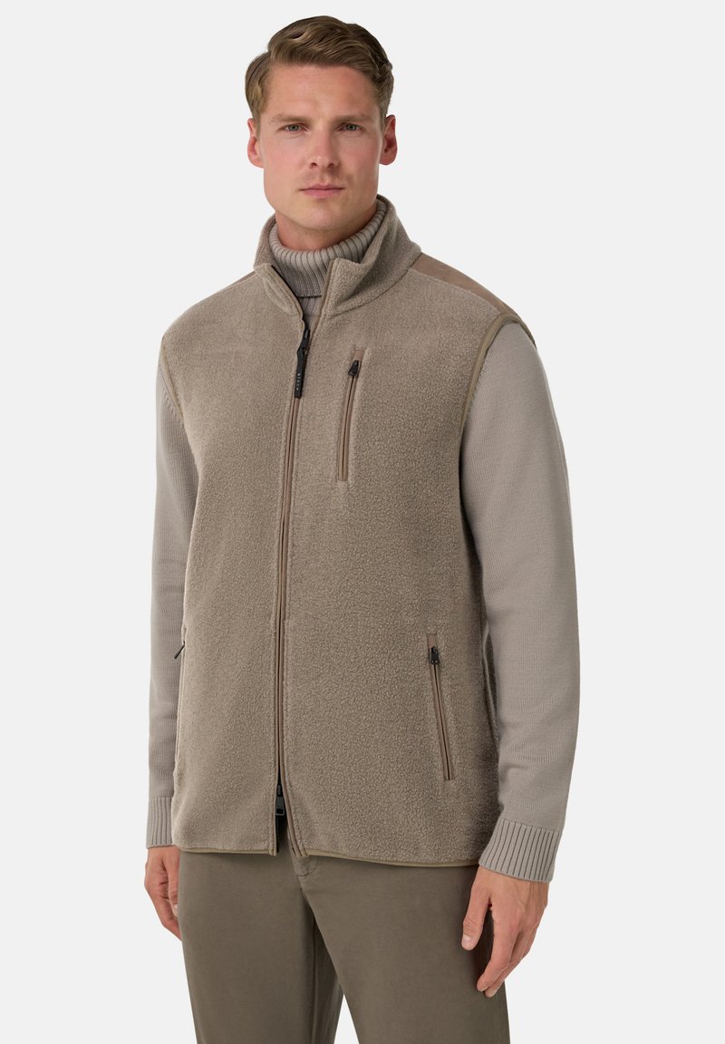 Mees, kes kannab beezist fliisvesti, millel on lukuga taskud, rõõmustades heledast pruunist kõrge kaelusega kampsunist ja sellele sobivatest pükstest, seistes ühtlase tausta ees.