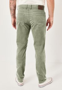 Grüne Babycord-Hosen mit schmaler Passform, ausgestattet mit zwei Gesäßtaschen und einem Lederpatch am Bund. Getragen mit weißen Sneakern.