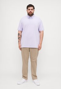 Ανοιχτό μωβ πόλο μπλουζάκι με γιακά και κοντά μανίκια, φορεμένο με μπεζ παντελόνι και λευκά αθλητικά παπούτσια. Περιέχει κεντητό λογότυπο στο στήθος.