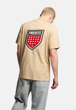 Osoba zwrócona tyłem, ubrana w beżowy T-shirt z czerwonym logo w kształcie tarczy przedstawiającym białe krzyże oraz napis "PROSTO.", połączony z jasnoniebieskimi jeansami.