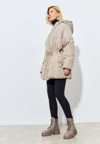 Chaqueta acolchada en color beige con cintura ajustable, capucha y bolsillos laterales; combinada con leggings negros y botas de tobillo marrones con cordones.