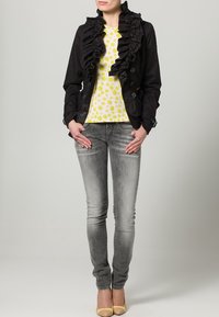 Svart jacka med rynkad framkant och knapprad, gul prickig skjorta, grå tajta jeans och gula klackskor. Jämn och slät textur över hela plaggen.