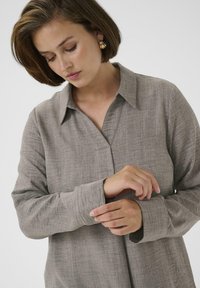 Blusa marrone, leggera, a maniche lunghe con colletto, davanti senza bottoni e con una sottile trama. Presenta maniche leggermente arrotolate.