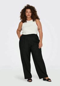 Γυναίκα με σγουρά μαλλιά στέκεται με το χέρι στην τσέπη, φορώντας λευκή αμάνικη μπλούζα, μαύρους φαρδιούς παντελόνες και μαύρες ψηλοτάκουνες σαγιονάρες.