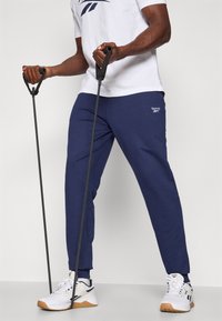 Námornícky športové nohavice so zúženými nohami, vyrobené z mäkkej tkaniny, majú logo Reebok. Model drží cvičebné odporové gumy a má na sebe biele tenisky.
