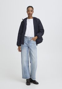 Pernata mornarsko plava jakna s kapuljačom, bijela majica i svijetloplave široke hlače. Model nosi smeđe gležnjače. Jednostavan, funkcionalan dizajn.