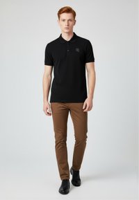 Černé polo tričko s tříknoflíkovou légou a jemným logem; kombinováno s hnědými slim-fit kalhotami a černými botami.