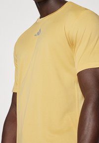 Żółta krótko rękawowa koszulka sportowa, teksturowany materiał, z małym szarym logo na klatce piersiowej, lekko luźny krój i okrągły dekolt.