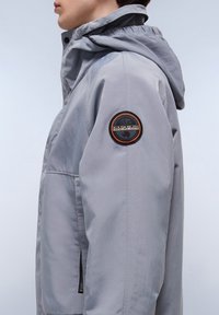 Gray hooded jacket made of lightweight fabric with a zippered pocket, featuring a circular brand patch on the left sleeve.